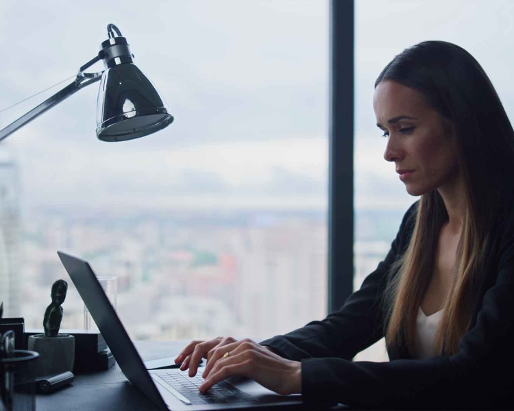 Eine Frau tippt auf ihrem Notebook am Schreibtisch. Durch das Fenster im Hintergrund sieht man Häuser einer Stadt.