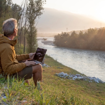 Mann sitzt am Fluss mit einem Laptop. Er blickt in die Ferne.
