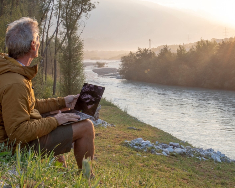 Mann sitzt am Fluss mit einem Laptop. Er blickt in die Ferne.