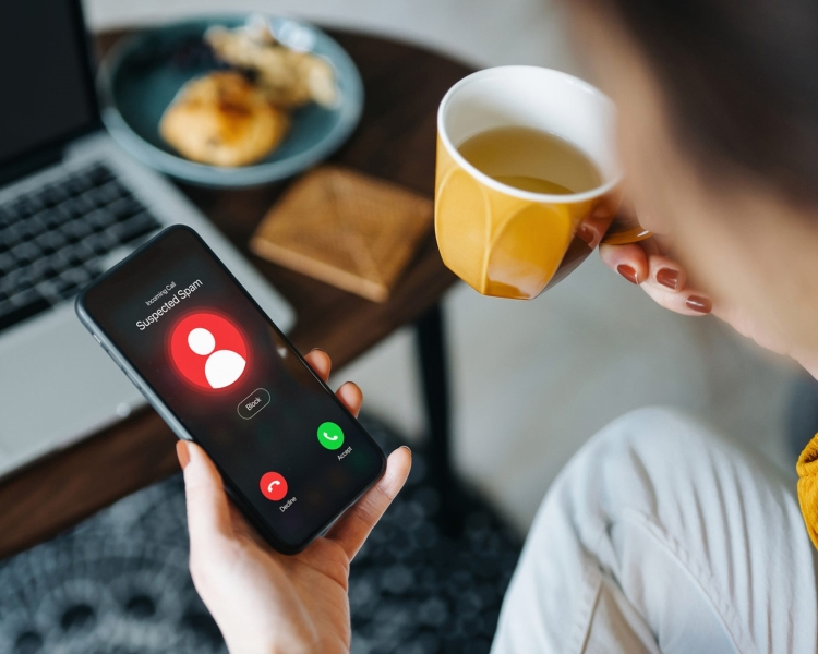 Eine Frau blickt auf ihr Smartphone, da eine unbekannte Nummer anruft und hält eine Tasse mit Kaffee in der anderen Hand