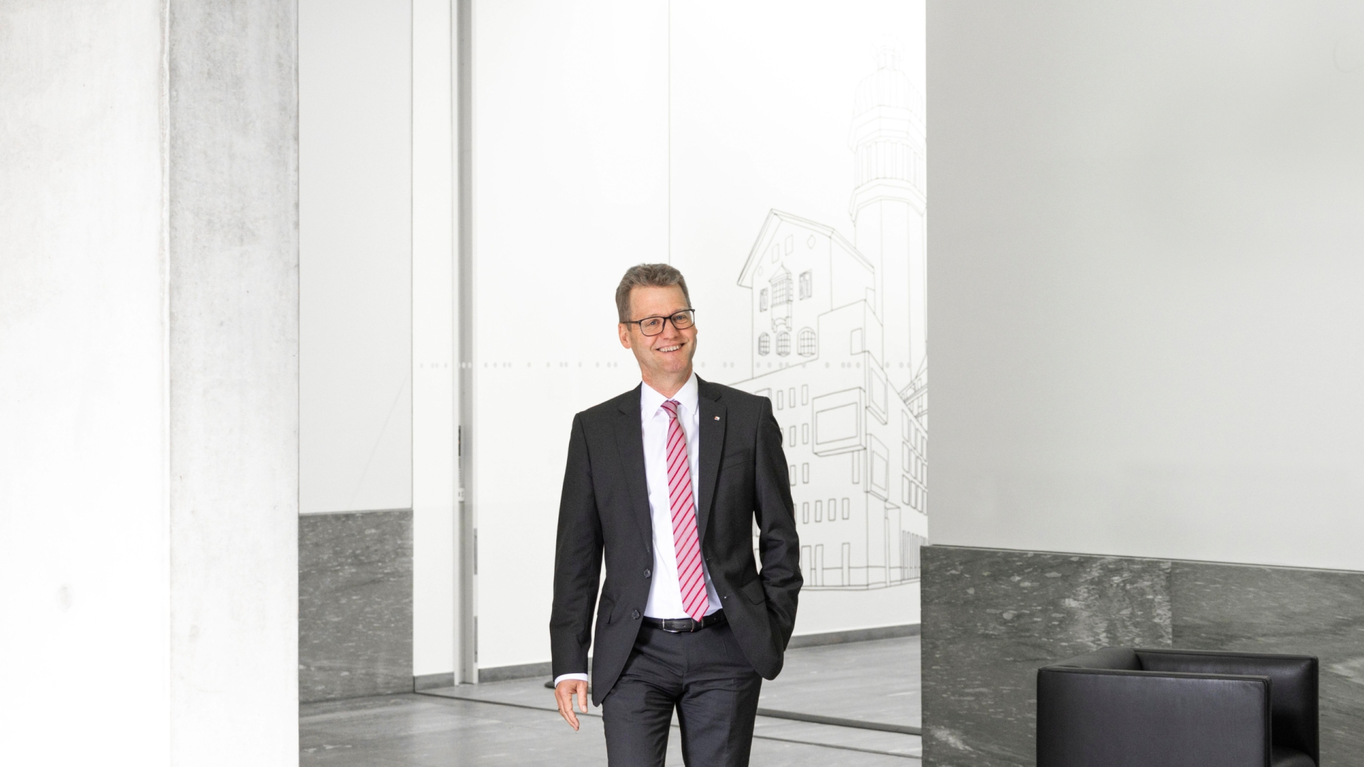 Ein Mann mit einer Brille trägt einen grauen Anzug inklusive rot-weißer Krawatte