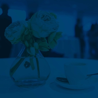 Espressotasse, -löffel und Unterteller auf einem Tisch mit weißer Tischdecke, daneben stehen Blumen in einer Vase, im Hintergrund sind Businessleute zu sehen, die sich unterhalten ( in Blau unterlegt)