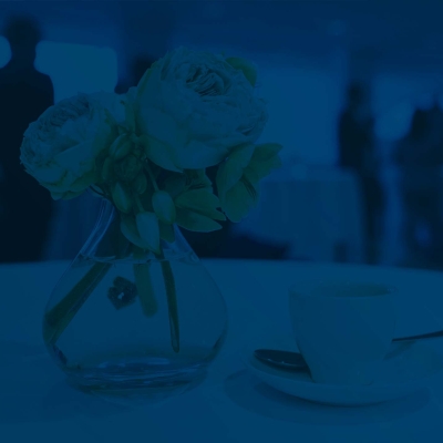 Espressotasse, -löffel und Unterteller auf einem Tisch mit weißer Tischdecke, daneben stehen Blumen in einer Vase, im Hintergrund sind Businessleute zu sehen, die sich unterhalten ( in Blau unterlegt)