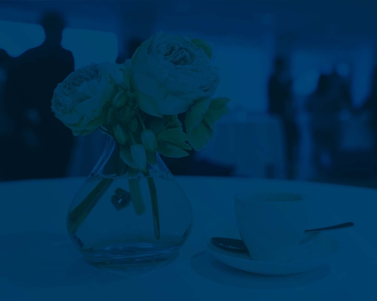 Espressotasse, -löffel und Unterteller auf einem Tisch mit weißer Tischdecke, daneben stehen Blumen in einer Vase, im Hintergrund sind Businessleute zu sehen, die sich unterhalten ( in Blau unterlegt)
