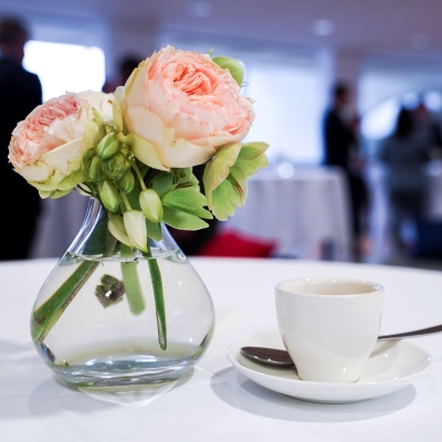 Espressotasse, -löffel und Unterteller auf einem Tisch mit weißer Tischdecke, daneben stehen Blumen in einer Vase, im Hintergrund sind Businessleute zu sehen, die sich unterhalten
