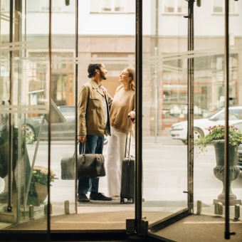Zwei Personen stehen mit Gepäck vor dem Eingang eines Stadthotels