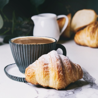 Eine Tasse Kaffee, eine Milchkanne und frisch gebackene Croissants auf einem Tisch, dahinter befindet sich eine grüne Pflanze