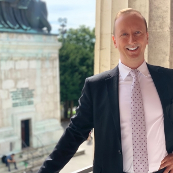 Matthias Weidmann mit Anzug und Krawatte vor einer Säule, dahinter ist ein Teil einer Kirche zu sehen, wo zwei Personen auf der Treppe sitzen