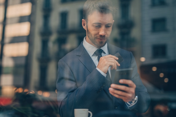 Ein Mann im Anzug sitzt am Tisch bei einer Tasse Kaffee und hält sein Smartphone in der linken sowie eine Kreditkarte in der rechten Hand