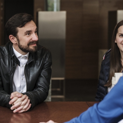 Zwei Personen stehen vor einem Bank Schalter und sprechen mit einem Mitarbeiter der Bank.