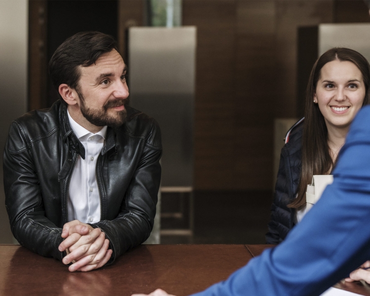 Zwei Personen stehen vor einem Bank Schalter und sprechen mit einem Mitarbeiter der Bank.