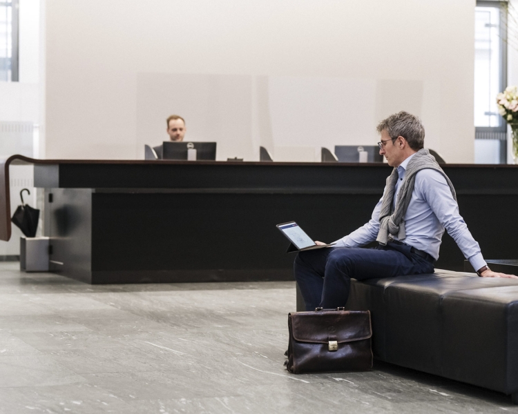 Ein Mann mit Brille sitzt auf einem Sessel in einer Bankfiliale und schaut auf seinem Tablet.
