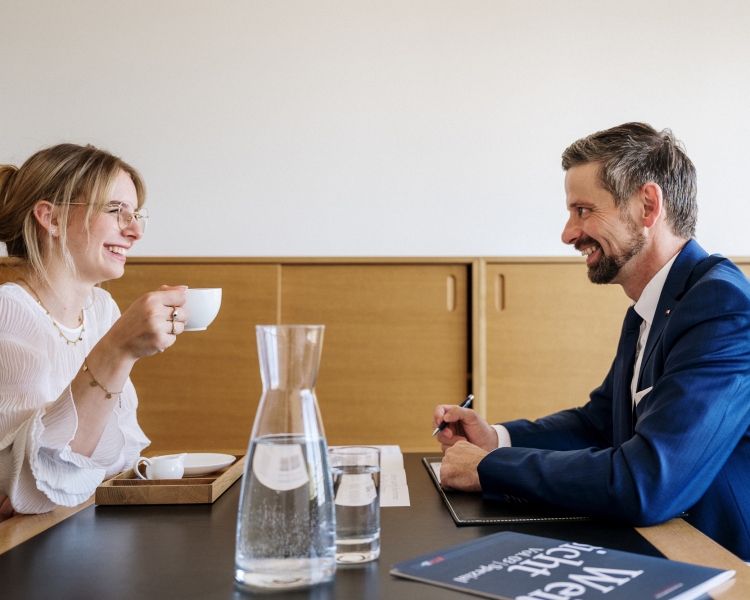 Zwei Personen unterhalten sich und lachen. Eine Person hält ein Tasse in der Hand.