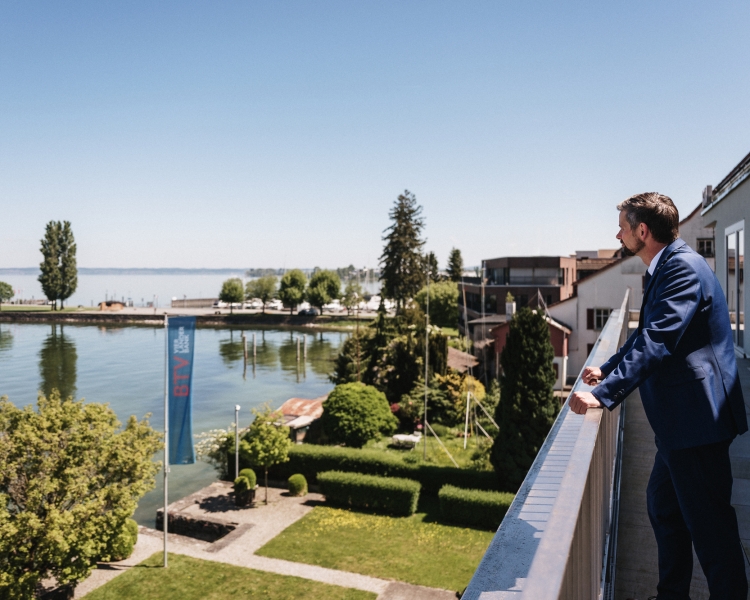 Ein Mann in Anzug steht auf einem Balkon und schaut auf einem See.