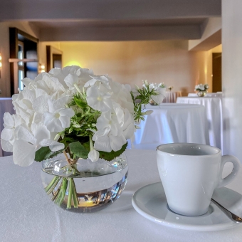Kaffeetasse, -löffel und Unterteller auf einem Tisch mit weißer Tischdecke, daneben stehen weiße Blumen in einer Vase, im Hintergrund sind weitere Stehtische zu sehen
