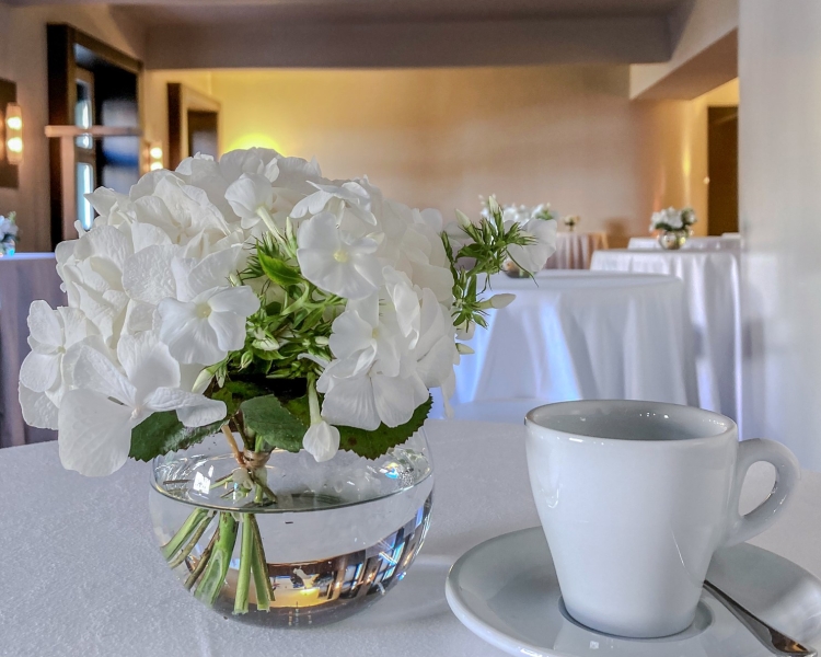 Kaffeetasse, -löffel und Unterteller auf einem Tisch mit weißer Tischdecke, daneben stehen weiße Blumen in einer Vase, im Hintergrund sind weitere Stehtische zu sehen
