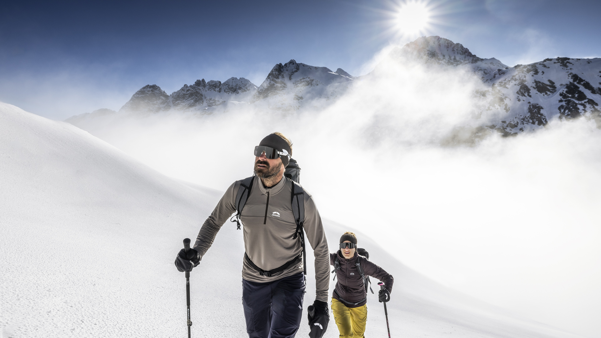 Ein Mann und eine Frau in Wanderbekleidung gehen auf den Berg umgeben von verschneiter Winterlandschaft und der Sonne im Rücken