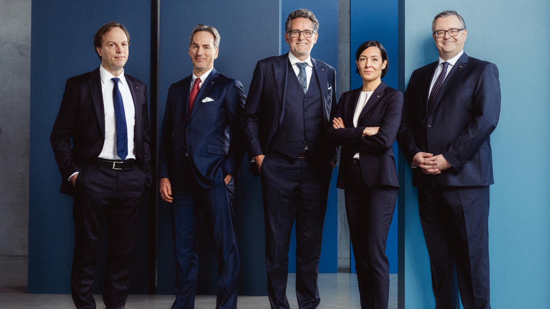 4 men and 1 woman are wearing a suit with a blue wall in the background.