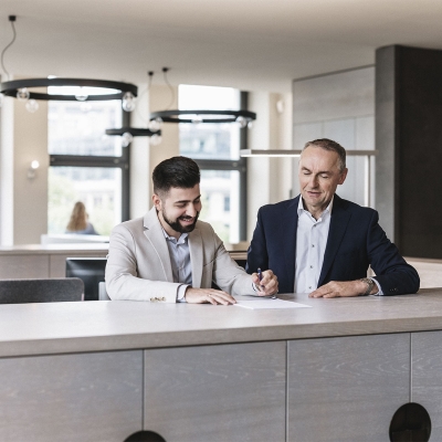 A man signs a document while standing next to another man.