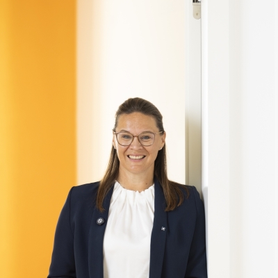 Claudia Polka, Gruppenleiterin Bankrechnungswesen der BTV Klagenfurt, trägt einen blauen Blazer und steht vor einer gelben Wand.
