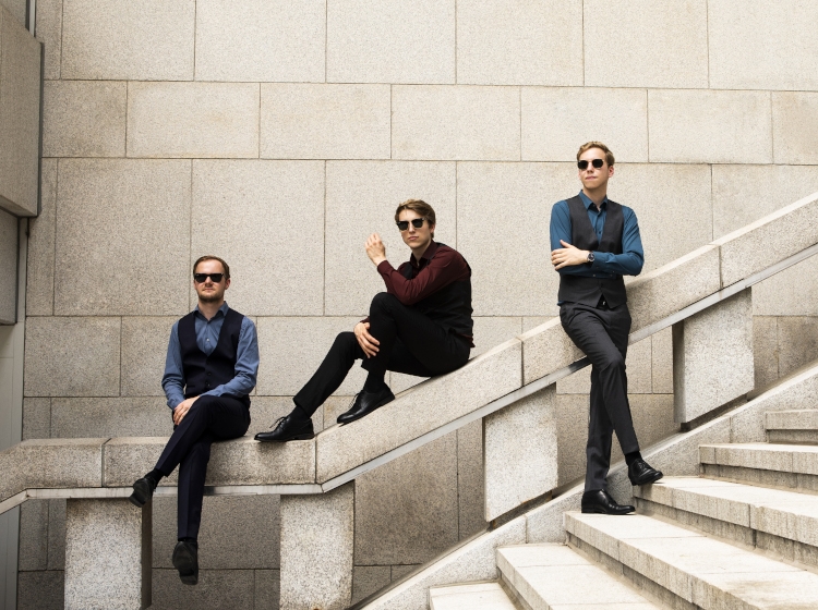 Drei Personen mit Sonnenbrille sind abgebildet. Zwei davon sitzen auf einem sandsteinfarbenen Treppengeländer, eine lehnt dagegen.