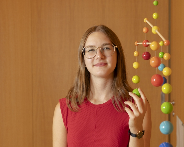 MUKKI‑Mitarbeiterin Sarah Bischofer steht vor der Kamera und lächelt, während sie ein Objekt in der Hand hält.