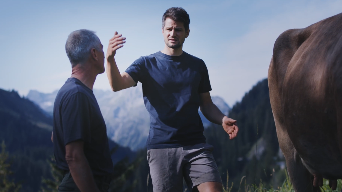 Skirennfahrer Johannes Strolz erklärt einen Publikum etwas. Im Hintergrund ist eine Alpenlandschaft zu sehen.