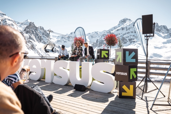 Drei Personen diskutieren ein Thema beim versus Festival, im Hintergrund ist eine Alpinlandschaft zu sehen.