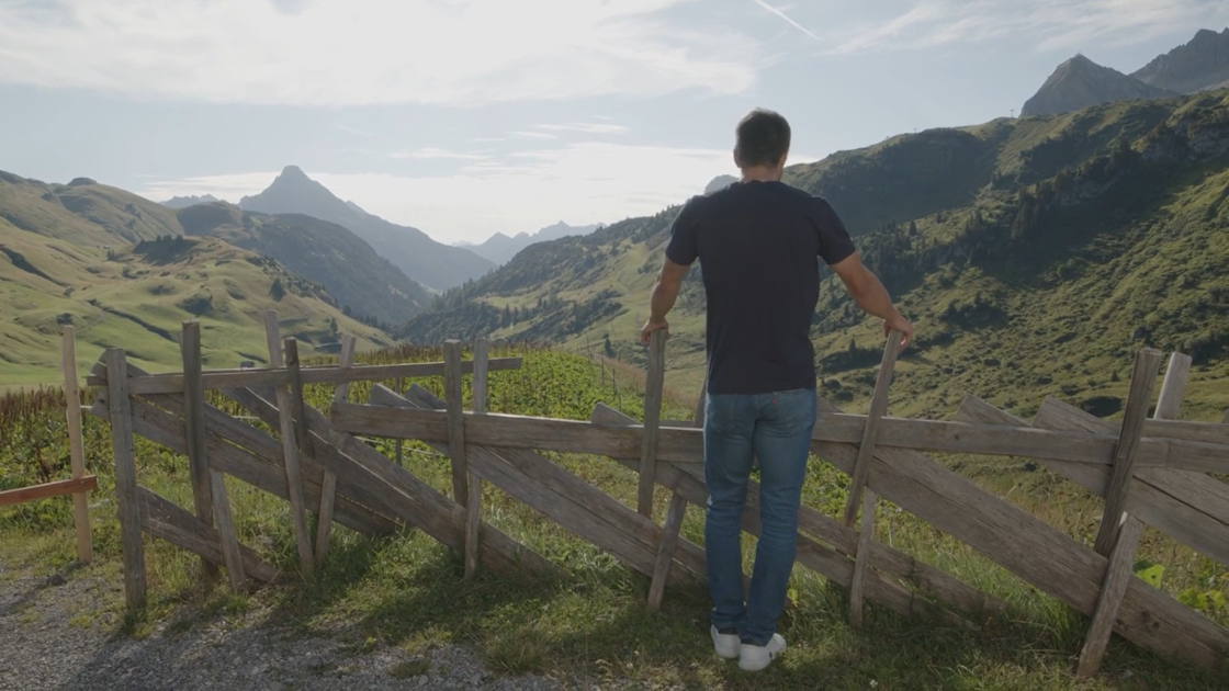 Skirennfahrer Johannes Strolz lehnt an einem Holzzaun und blickt in ein weites Alpental mit Bergen und Wiesen.