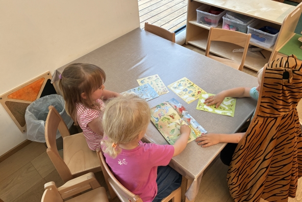 Kinder der MUKKI‑Kinderkrippe kleben gemeinsam Aufkleber in ein Klebebuch und gestalten konzentriert ihre Seiten.