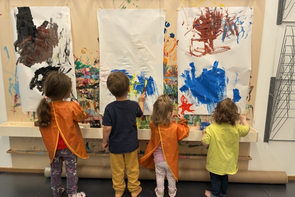 Kinder der MUKKI‑Kinderkrippe malen gemeinsam mit Farbe auf mehreren Papierstücken und gestalten farbenfrohe Bilder an einer Wand.