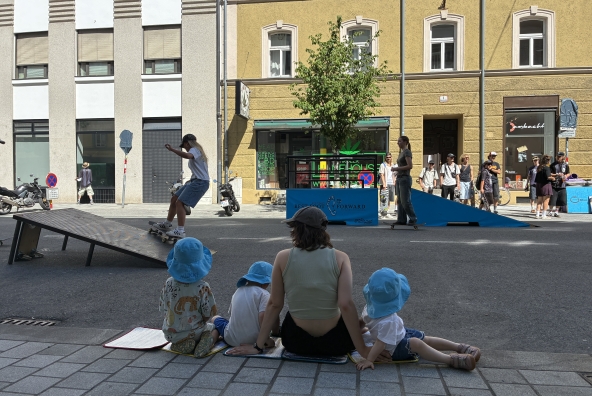 Kinder eine Begleitperson der MUKKI‑Kinderkrippe beobachten gemeinsam eine Skaterin in Innsbruck und verfolgen interessiert ihre Bewegungen.