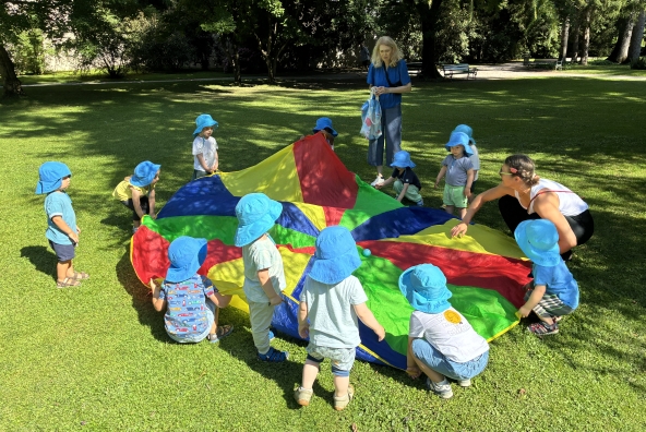 Kinder der MUKKI‑Kinderkrippe spielen gemeinsam in einem Park und halten eine große Decke fest, während sie miteinander interagieren.