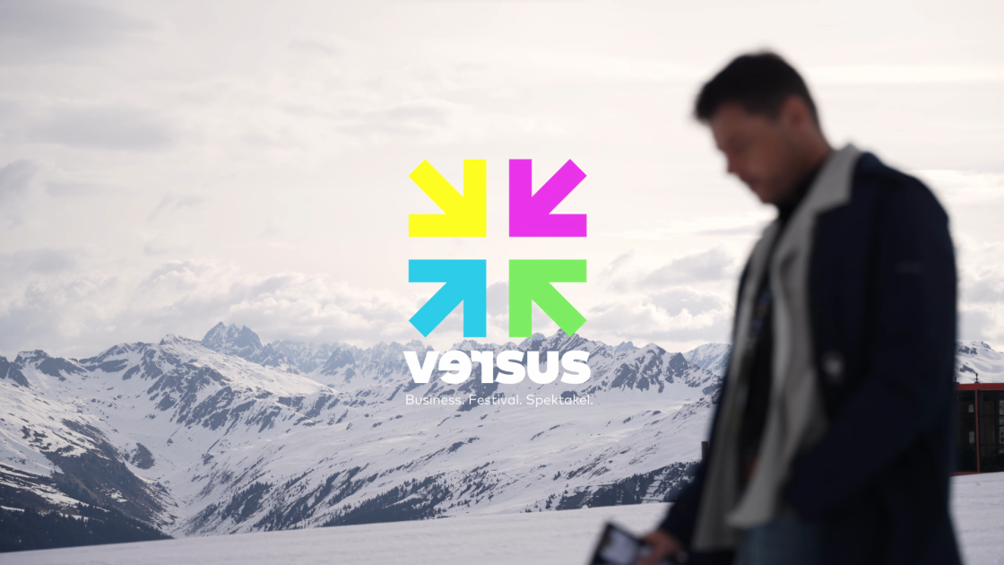 Alpine Berglandschaft mit dem Versus‑Festival‑Logo; im Vordergrund steht ein Mann und und läuft vorbei.