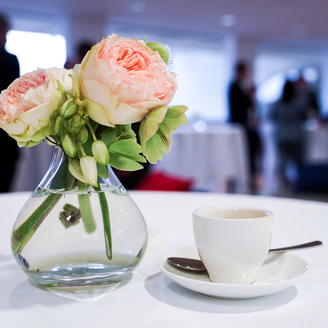 Espressotasse, -löffel und Unterteller auf einem Tisch mit weißer Tischdecke, daneben stehen Blumen in einer Vase, im Hintergrund sind Businessleute zu sehen, die sich unterhalten