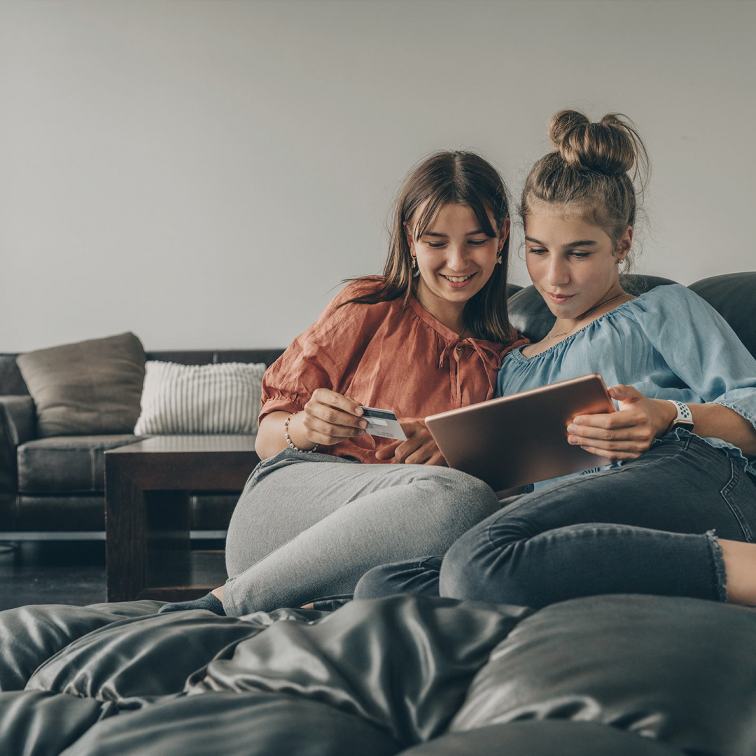 Zwei Freundinnen sitzen auf der Couch und bezahlen mit einer Debitkarte im Internet