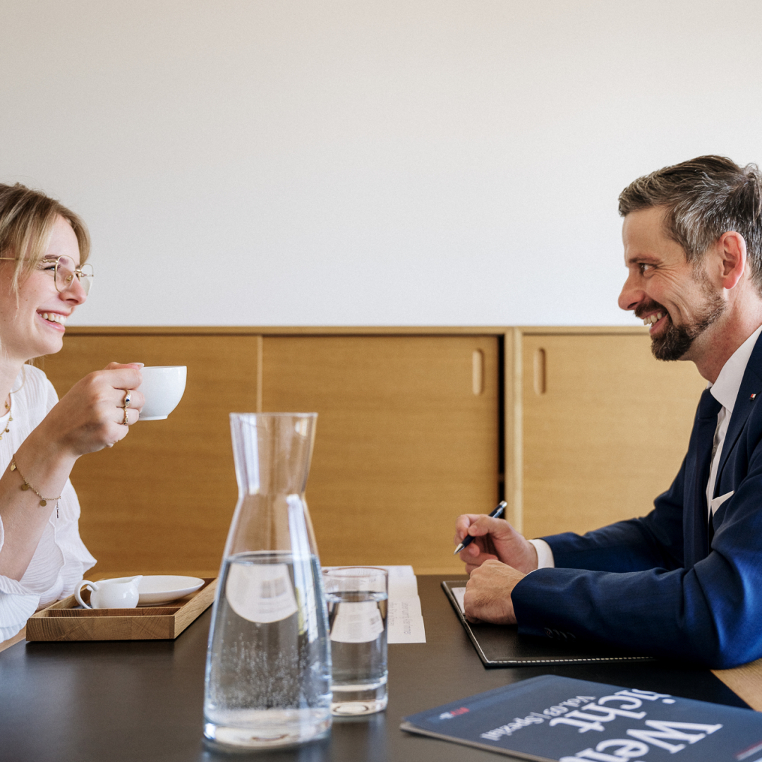 Zwei Personen unterhalten sich und lachen. Eine Person hält ein Tasse in der Hand.