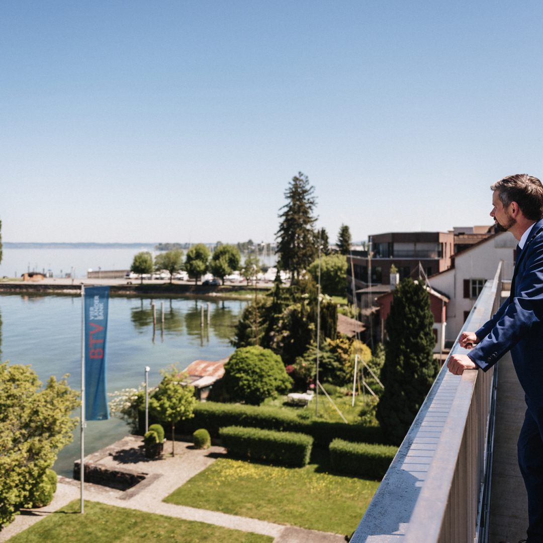 Ein Mann in Anzug steht auf einem Balkon und schaut auf einem See.